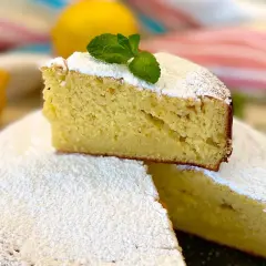 Una fetta di torta allo yogurt greco, soffice e spolverata di zucchero a velo, decorata con menta fresca. Due limoni sullo sfondo.
