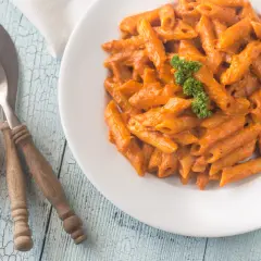 Piatto di penne alla vodka cremose, guarnite con prezzemolo, su un tavolo di legno azzurro. Pomodorini freschi e posate completano la scena.