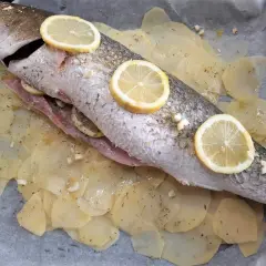 Ombrina al forno pronta per essere infornata, adagiata su patate affettate e guarnita con fette di limone e aglio.