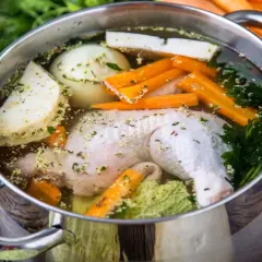 Pentola con brodo di pollo, verdure fresche e erbe aromatiche. Carote intere sullo sfondo.