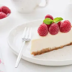 Una fetta di torta fredda allo yogurt, guarnita con lamponi freschi e menta, su un piatto bianco con una forchetta.