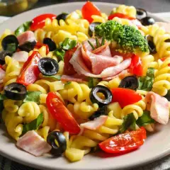 Un piatto di pasta fredda saporita con fusilli, pomodorini, olive nere, prosciutto cotto e un ciuffo di broccoli.