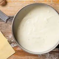 Besciamella ricetta: una cremosa salsa bianca in pentola, con sfoglie di pasta, noce moscata e grattugia su un tavolo di legno.