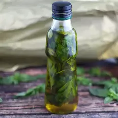 Bottiglia di vetro piena di olio al basilico, con foglie fresche sparse sul tavolo di legno.