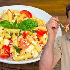 Un uomo sorride mentre assaggia una forchettata di pasta alla crudaiola, un piatto fresco con pomodorini, mozzarella e basilico.