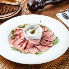 Fettine di arrosto rosa disposte a cerchio su un piatto bianco, con una ciotolina di salsa tonnata guarnita da un cappero.