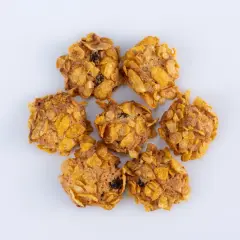 Biscotti croccanti con fiocchi di mais e uvetta, simili a rose del deserto, disposti a cerchio su sfondo bianco.