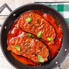 Melanzane in padella, succulente e cotte in un ricco sugo di pomodoro, guarnite con foglie di basilico fresco.