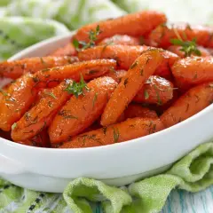 Carote glassate in padella, profumate con erbe aromatiche, servite in una ciotola bianca su un tovagliolo verde.