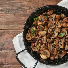 Funghi in padella, dorati e profumati, guarniti con prezzemolo fresco, pronti per essere gustati su un tavolo di legno rustico.