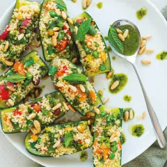 Zucchine ripiene di cous cous colorato con verdure, pinoli e menta, servite su un piatto bianco con un cucchiaio di salsa verde.