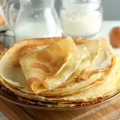 Pila di crepes dorate, pronte per essere farcite. Ingredienti per la ricetta crepes visibili sullo sfondo: uova, latte e farina.