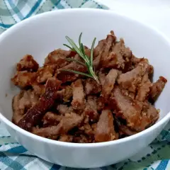 Deliziosi straccetti di vitello cotti e guarniti con un rametto di rosmarino, serviti in una ciotola bianca su un tovagliolo a quadri.