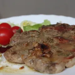 Una succulenta braciola di maiale, condita con erbe aromatiche, servita con pomodorini e insalata fresca su un piatto bianco.