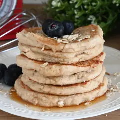 Pila di pancake con farina di avena, guarniti con mirtilli e sciroppo d'acero. Sullo sfondo, una bilancia rossa e un frullino.