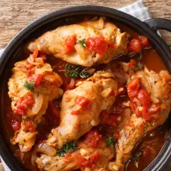 Coniglio all'ischitana in tegame, con pomodoro, cipolla e erbe aromatiche, servito su un tavolo di legno.