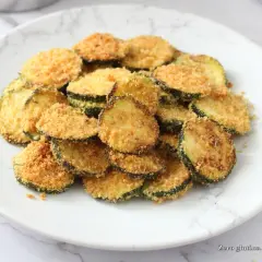 Fettine di zucchine impanate e cotte nella friggitrice ad aria, dorate e croccanti, servite su un piatto bianco.
