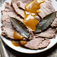 Fette di brasato di manzo succulento su un piatto, nappate con una salsa dorata e guarnite con foglie di alloro.