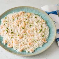 Un cremoso risotto gamberi e limone, guarnito con erba cipollina, servito su un piatto turchese. Accanto, un tovagliolo con pesci blu e una forchetta.