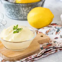 Una ciotola di cremosa maionese fatta in casa, guarnita con erbe fresche, accanto a limoni, un uovo e una frusta.