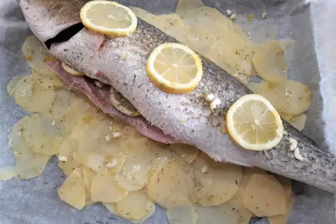 Ombrina al forno pronta per essere infornata, adagiata su patate affettate e guarnita con fette di limone e aglio.