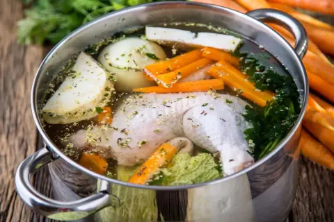Pentola con brodo di pollo, verdure fresche e erbe aromatiche. Carote intere sullo sfondo.