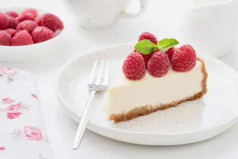 Una fetta di torta fredda allo yogurt, guarnita con lamponi freschi e menta, su un piatto bianco con una forchetta.