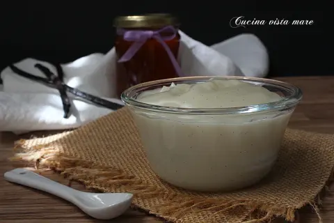 Una ciotola di cremosa crema al latte, profumata alla vaniglia, pronta per essere gustata. Accanto, un barattolo di marmellata e bacche di vaniglia.