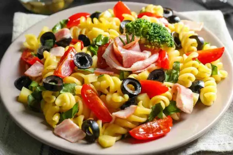 Un piatto di pasta fredda saporita con fusilli, pomodorini, olive nere, prosciutto cotto e un ciuffo di broccoli.