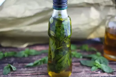 Bottiglia di vetro piena di olio al basilico, con foglie fresche sparse sul tavolo di legno.