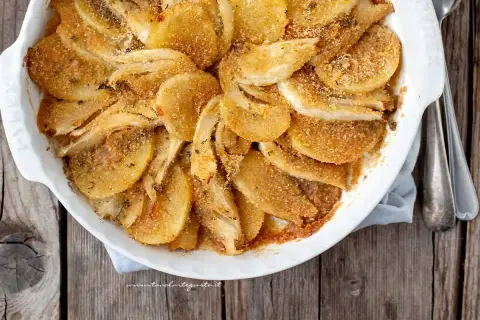 Finoçchi gratinati al forno, dorati e croccanti, in una pirofila bianca su un tavolo di legno.