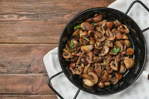 Funghi in padella, dorati e profumati, guarniti con prezzemolo fresco, pronti per essere gustati su un tavolo di legno rustico.