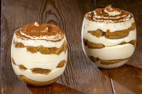 Due bicchieri di tiramisù senza uova, stratificati con crema e savoiardi, spolverati di cacao.