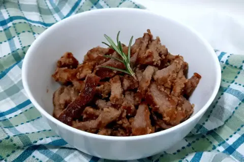 Deliziosi straccetti di vitello cotti e guarniti con un rametto di rosmarino, serviti in una ciotola bianca su un tovagliolo a quadri.