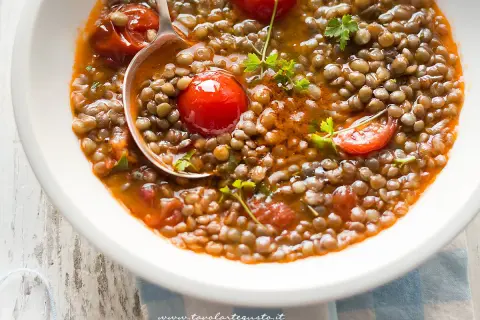 Una ciotola di zuppa di lenticchie fumante, con pomodorini e prezzemolo fresco. Un cucchiaio serve una porzione generosa di questo piatto confortante.