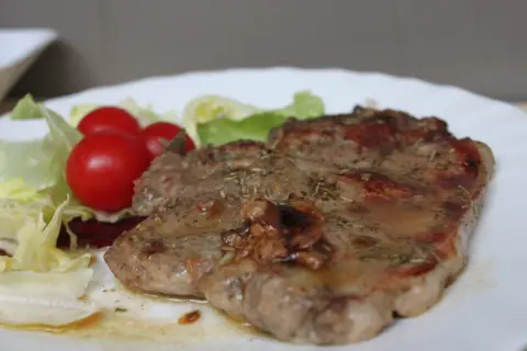 Una succulenta braciola di maiale, condita con erbe aromatiche, servita con pomodorini e insalata fresca su un piatto bianco.