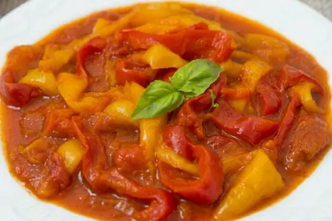 Peperonata ricetta della nonna: peperoni rossi e gialli stufati in un sugo saporito, guarniti con una fogliolina di basilico.