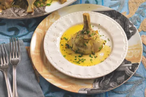Un carciofo alla romana, tenero e profumato, servito su un piatto bianco con olio e prezzemolo.