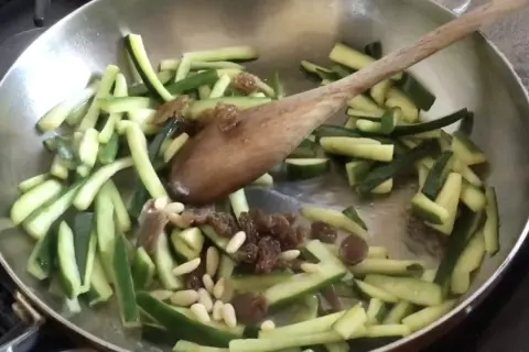 Zucchine tagliate a julienne con uvetta e pinoli in padella, pronte per essere trasformate in un delizioso piatto agrodolce.