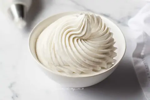 Una ciotola bianca piena di soffice crema chantilly, con striature perfette e puntini di vaniglia visibili.