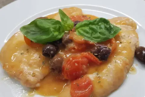 Delizioso pollo alla mediterranea con pomodorini, olive nere e basilico fresco su un piatto bianco.