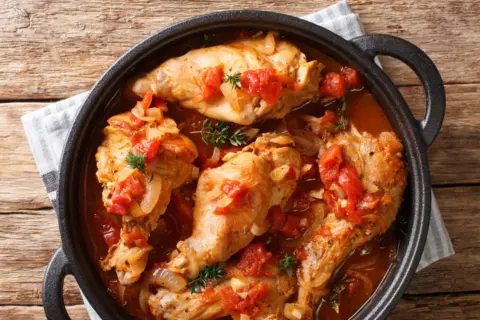 Coniglio all'ischitana in tegame, con pomodoro, cipolla e erbe aromatiche, servito su un tavolo di legno.