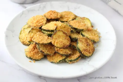 Fettine di zucchine impanate e cotte nella friggitrice ad aria, dorate e croccanti, servite su un piatto bianco.