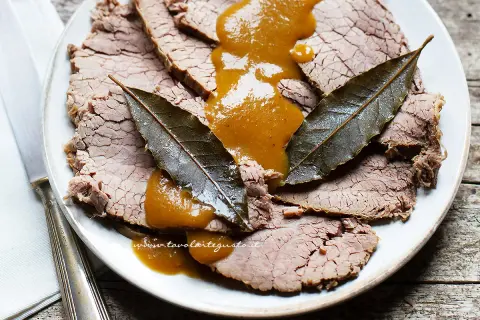 Fette di brasato di manzo succulento su un piatto, nappate con una salsa dorata e guarnite con foglie di alloro.