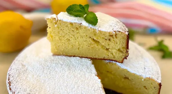 Una fetta di torta allo yogurt greco, soffice e spolverata di zucchero a velo, decorata con menta fresca. Due limoni sullo sfondo.