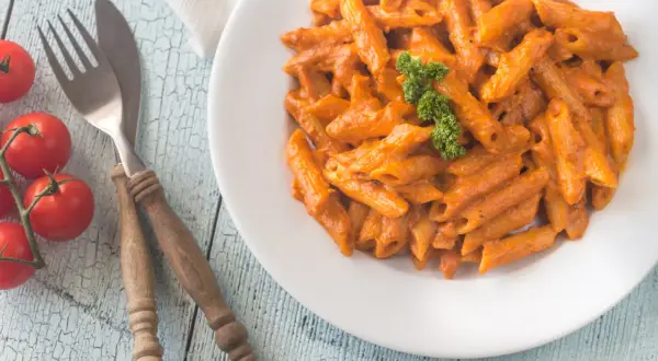 Piatto di penne alla vodka cremose, guarnite con prezzemolo, su un tavolo di legno azzurro. Pomodorini freschi e posate completano la scena.