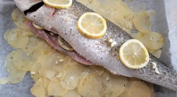 Ombrina al forno pronta per essere infornata, adagiata su patate affettate e guarnita con fette di limone e aglio.