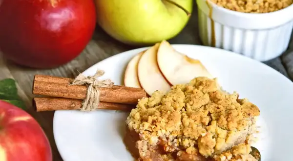 Un delizioso crumble di mele, con mele rosse e verdi sullo sfondo, cannella e fette di mela.