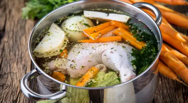 Pentola con brodo di pollo, verdure fresche e erbe aromatiche. Carote intere sullo sfondo.