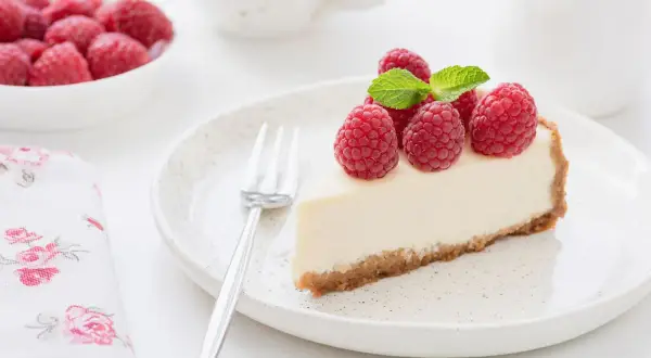 Una fetta di torta fredda allo yogurt, guarnita con lamponi freschi e menta, su un piatto bianco con una forchetta.